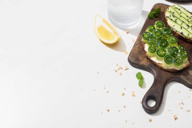 Taze salatalıklı tostlar ahşap kesim tahtasının üzerinde, suyun yanında ve limonlu beyaz arka planda