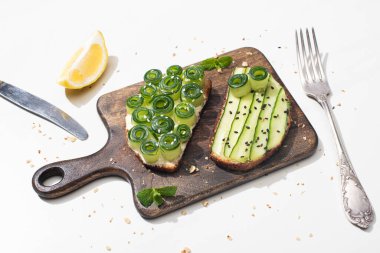 Taze salatalıklı tostlar ahşap kesim tahtasında çatal bıçak takımı yanında ve limonlu beyaz arka planda.