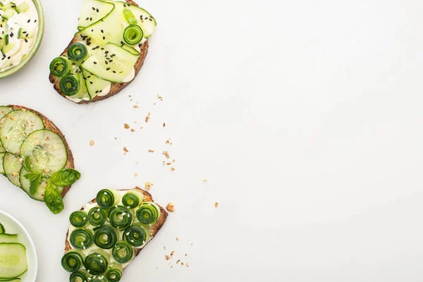 top view of fresh cucumber toasts with seeds, basil leaves near yogurt in bowl on white background