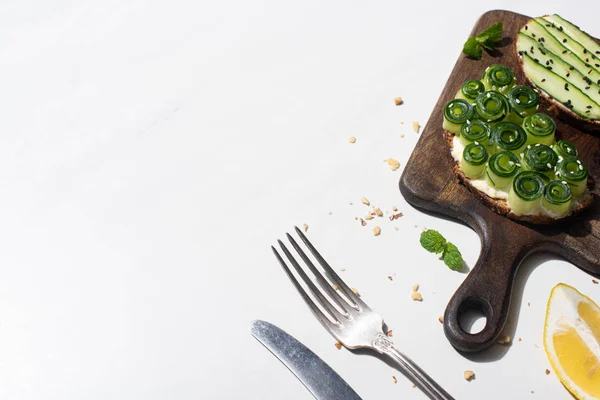 fresh cucumber toasts on wooden cutting board near cutlery and lemon on white background