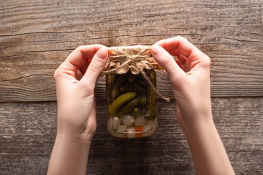 cropped view of woman holding bow on homemade tasty canned cucumbers in jar on wooden table clipart