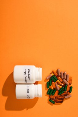Top view of containers with fish oil and dietary supplements lettering near colorful capsules on orange background clipart