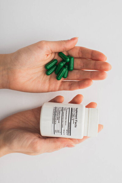 Cropped view of woman holding container and green capsules on white background 
