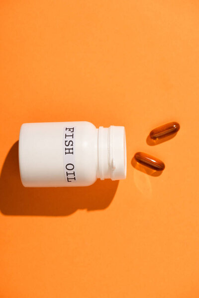 Вид сверху контейнера с рыбьим жиром, лежащим рядом с капсулами на оранжевом фоне
