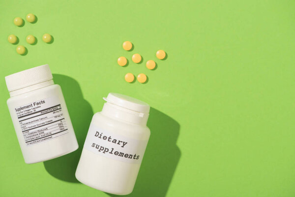 Top view of containers with dietary supplements lettering and pills on green background