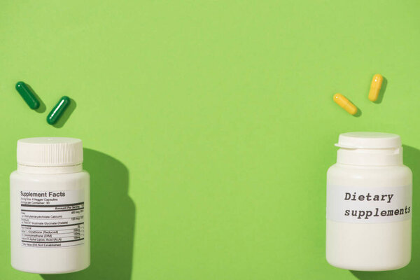 Top view of containers with dietary supplements lettering and colorful capsules on green background