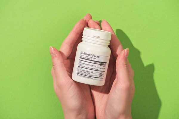 Cropped view of woman holding container with dietary supplements on green background