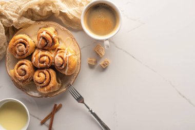top view of fresh homemade cinnamon rolls on marble surface with cup of coffee, condensed milk, fork and cloth clipart