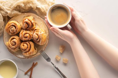 cropped view of woman holding cup of coffee near homemade cinnamon rolls on marble surface with fork, condensed milk and cloth clipart