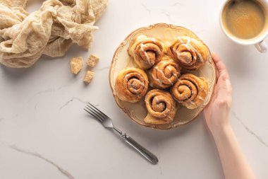 Ev yapımı tarçın rulolarını çatal, kahve ve kumaşla mermer yüzeyde tutan bir kadın.