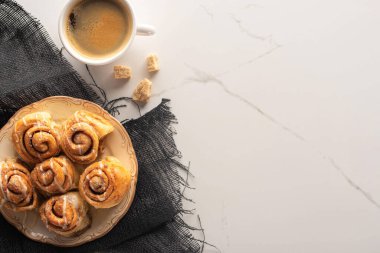 Taze ev yapımı tarçın rulolarının mermer yüzeyinde bir fincan kahve, kahverengi şeker ve kumaş ile üst görüntüsü