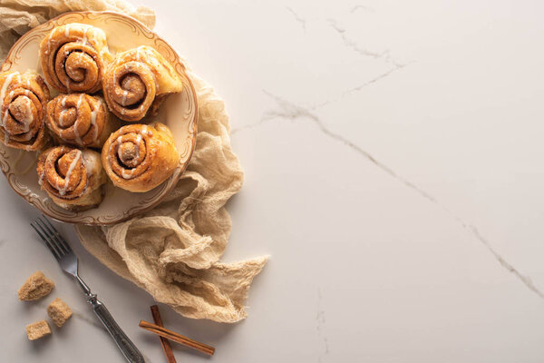 top view of fresh homemade cinnamon rolls on marble surface with brown sugar, cinnamon sticks, fork and cloth