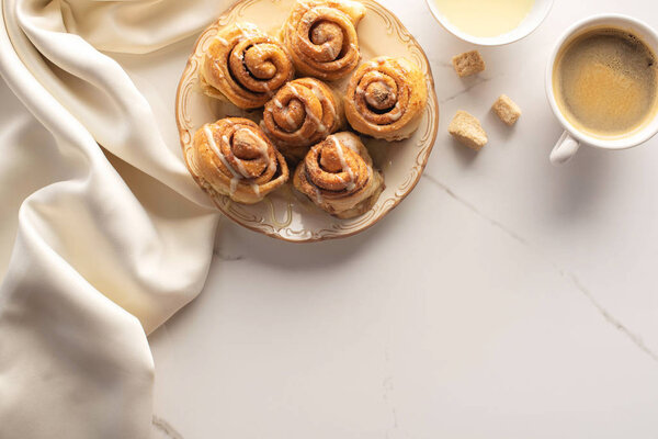top view of fresh homemade cinnamon rolls on marble surface with cup of coffee, brown sugar, condensed milk and silk cloth