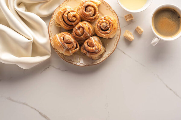 top view of fresh homemade cinnamon rolls on marble surface with cup of coffee, brown sugar, condensed milk and silk cloth