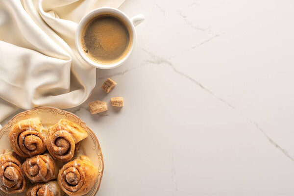 top view of fresh homemade cinnamon rolls on marble surface with cup of coffee, brown sugar and silk cloth
