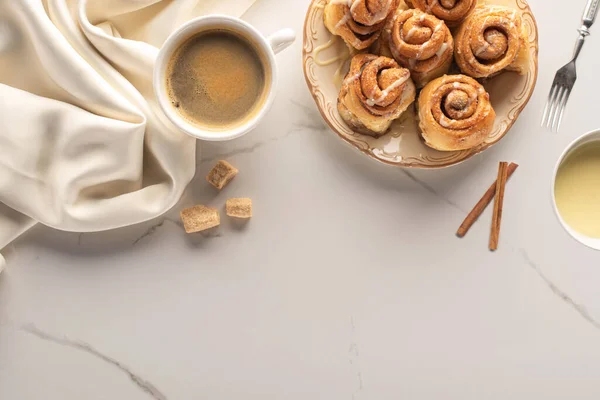 用咖啡 浓缩牛奶 叉子和丝布在大理石表面上制作的新鲜肉桂卷的顶部视图 — 图库照片