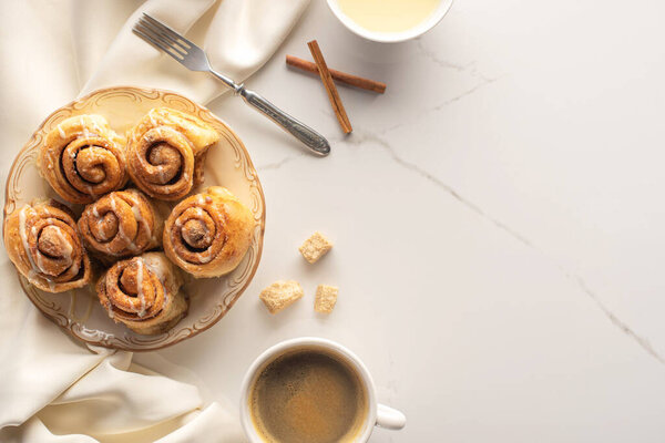 top view of fresh homemade cinnamon rolls on marble surface with coffee, condensed milk, fork and satin cloth