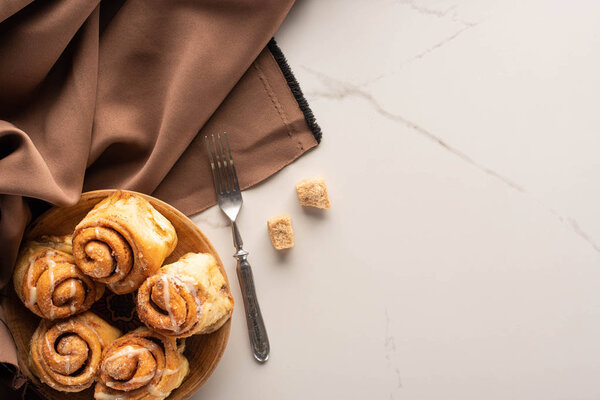 top view of fresh homemade cinnamon rolls on marble surface with brown sugar, fork and brown silk cloth