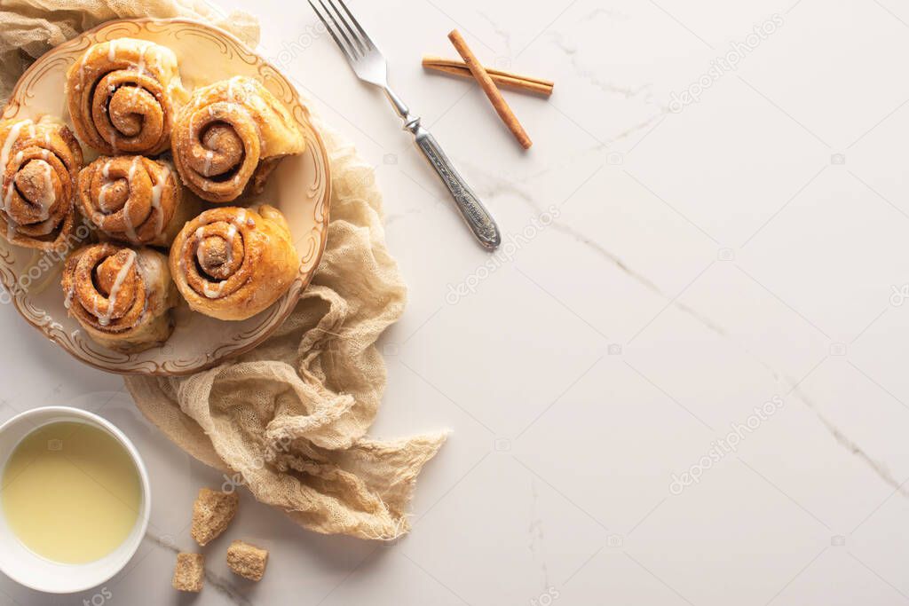top view of fresh homemade cinnamon rolls on marble surface with condensed milk, brown sugar, cinnamon sticks, fork and cloth