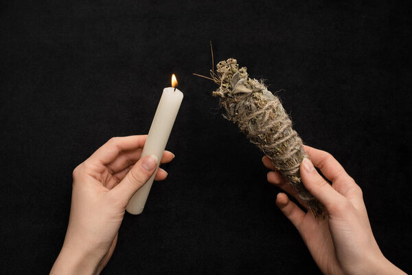 Cropped view of shaman holding burning candle and smudge stick isolated on black 