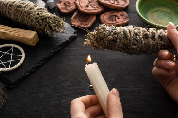 Cropped view of shaman holding candle and smudge stick near witchcraft on black wooden background
