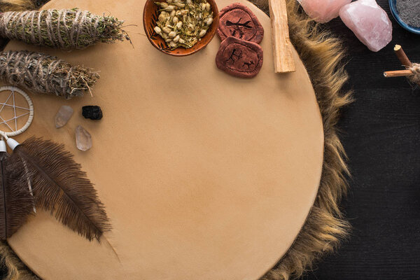 Top view of shamanic dreamcatcher, herbs and crystals on tambourine on black wooden surface