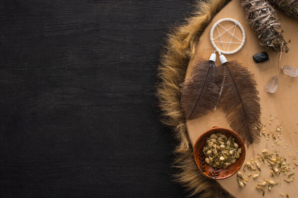 Top view of smudge sticks near dreamcatcher with feathers and crystals on shamanic tambourine on dark wooden surface