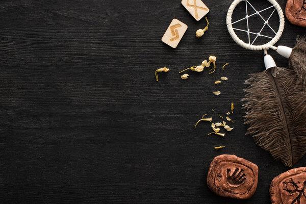 Top view of dreamcatcher, runes and dry flowers on black wooden background with copy space