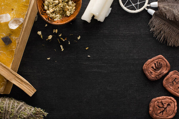 Top view of clay amulets, book with dreamcatcher and crystals on black wooden background