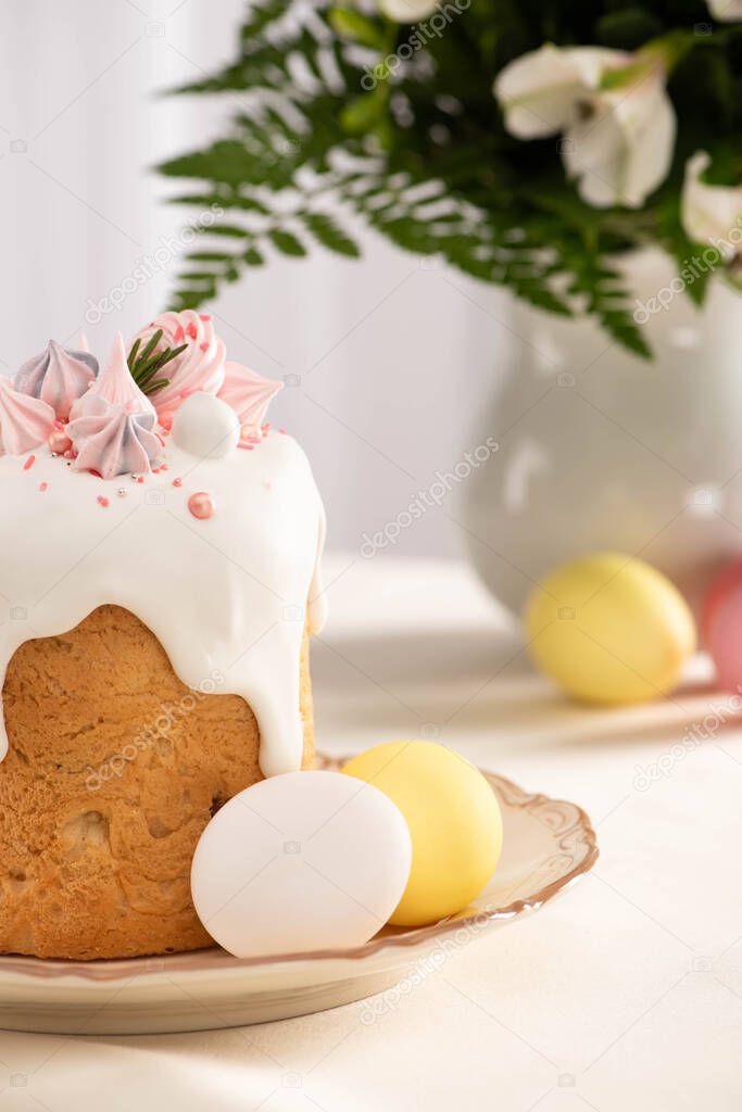 selective focus of delicious Easter cake decorated with meringue near colorful eggs on plate on table with vase of flowers