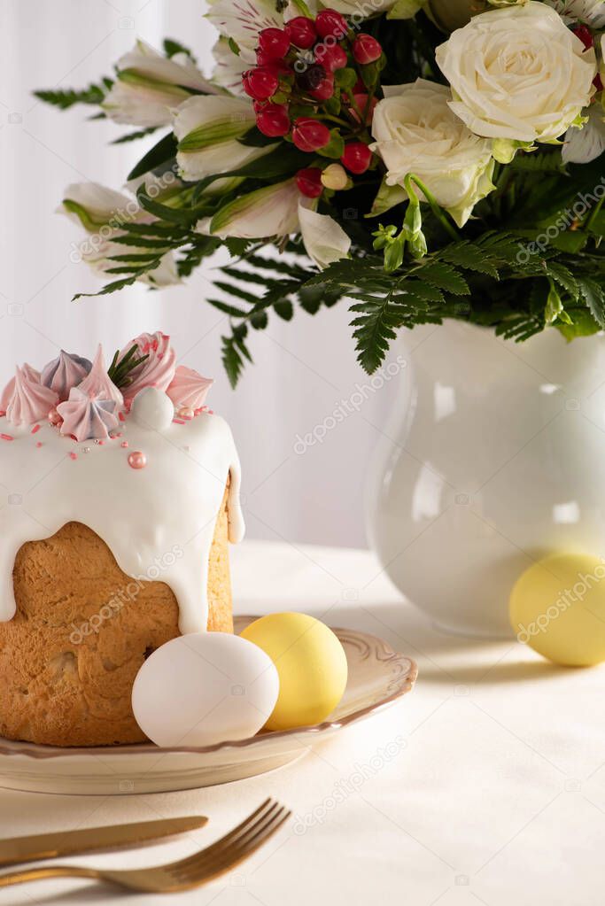delicious Easter cake decorated with meringue near colorful eggs on plate on table with vase of flowers and cutlery
