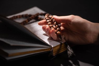 cropped view of woman holding rosary with cross near open holy bible in dark with sunlight clipart