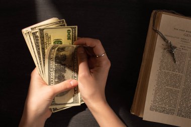 KYIV, UKRAINE - JANUARY 17, 2020: cropped view of woman counting money near holy bible with cross on dark background with sunlight