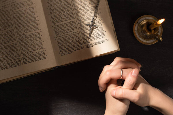 KYIV, UKRAINE - JANUARY 17, 2020: cropped view of woman praying near open holy bible with cross on dark background with sunlight