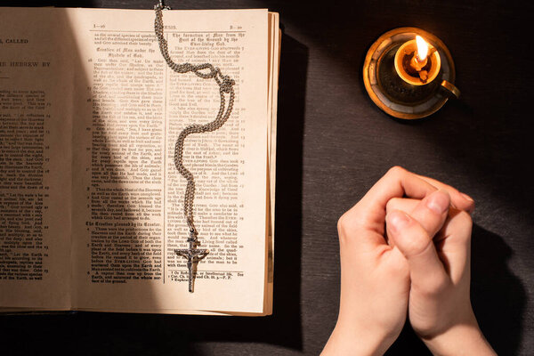 KYIV, UKRAINE - JANUARY 17, 2020: cropped view of woman praying near holy bible with cross, candle on dark background with sunlight