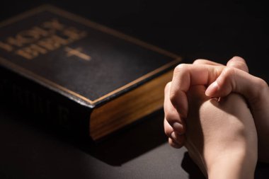 cropped view of woman praying near holy bible in dark clipart