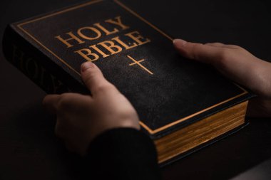 cropped view of woman holding holy bible in dark clipart