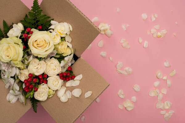 top view of bouquet of flowers in cardboard box near scattered petals on pink background