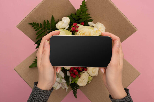 cropped view of woman holding smartphone above bouquet of flowers in cardboard box on pink background