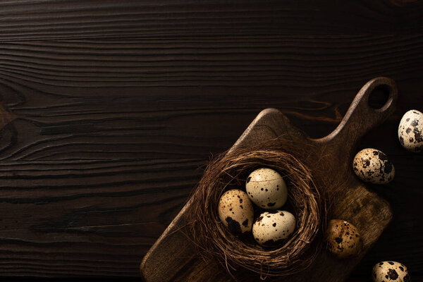 top view of quail eggs in nest on brown cutting board on dark wooden background