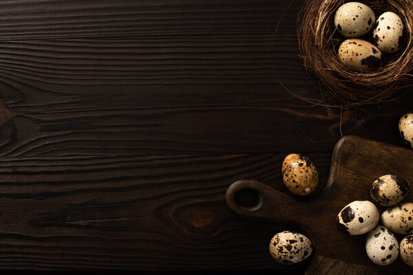 top view of quail eggs in brown nest and on chopping board on dark wooden background
