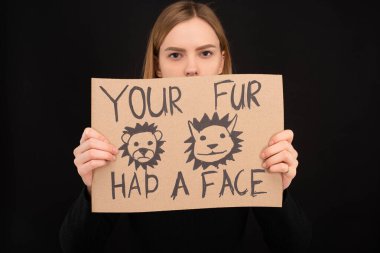 woman with obscure face holding cardboard sign with your fur had a face inscription isolated on black clipart