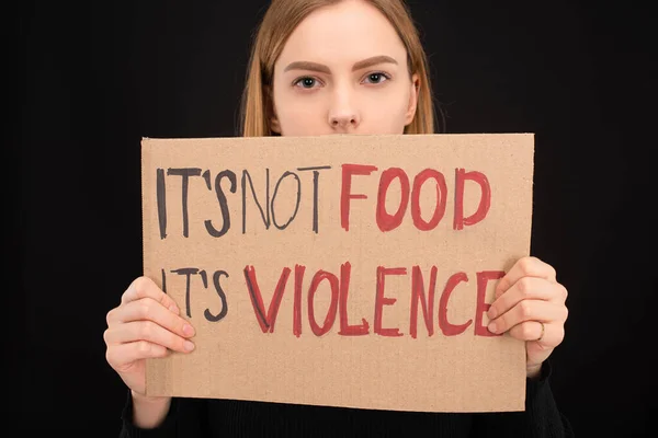woman with obscure face holding cardboard sign with its not food its violence inscription isolated on black