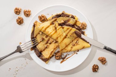 top view of tasty crepes with chocolate spread and walnuts on plate with cutlery on grey background clipart