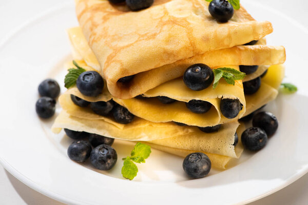 close up view of tasty crepes with mint and blueberries on plate on white background