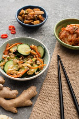 selective focus of bowls with tasty kimchi near chopsticks and ginger on concrete surface clipart