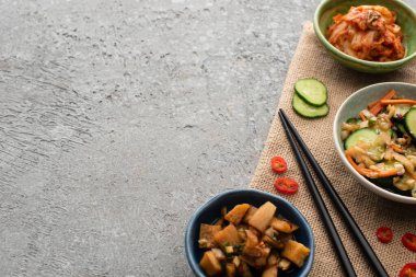 top view of bowls with tasty kimchi near chopsticks, sliced cucumber and chili pepper on sackcloth on concrete surface clipart