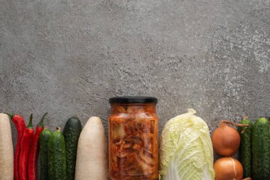 top view of chili peppers, cucumbers, daikon radish, chinese cabbage, onions and kimchi jar on grey concrete background clipart