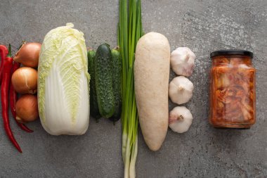 top view of chili peppers, kimchi jar, green onions, cucumbers, daikon radish, chinese cabbage, onions and garlic on grey concrete background clipart