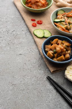selective focus of bowls with kimchi near ginger, sliced cucumber and chili pepper on sackcloth near chopsticks on concrete surface clipart